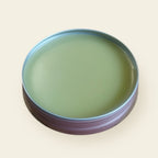 Open rose-gold tin of Melzayta Body Balm, smooth golden-green balm, on a cream background